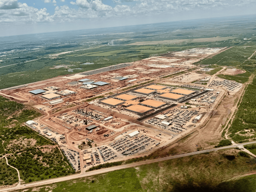 Aerial view of a massive data center construction site stretching across a large landscape, with rows of industrial buildings, construction areas, dirt roads, equipment, and cleared land surrounded by green fields and natural terrain under a cloudy sky.
