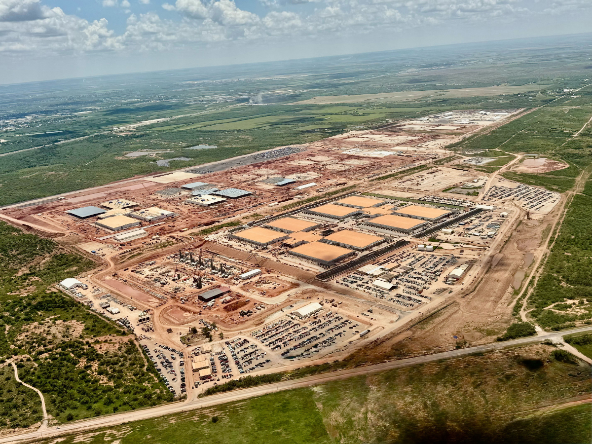 Aerial view of a massive data center construction site stretching across a large landscape, with rows of industrial buildings, construction areas, dirt roads, equipment, and cleared land surrounded by green fields and natural terrain under a cloudy sky.